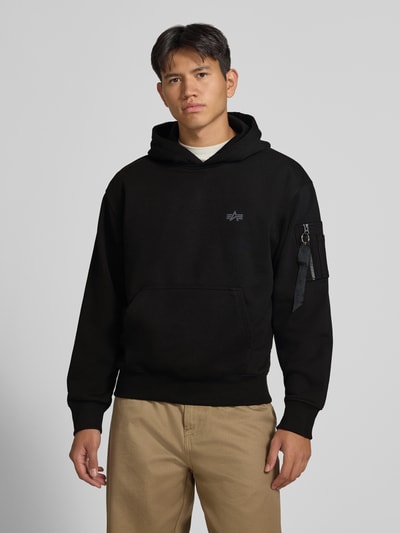 Männermodel trägt Hoodie mit Label-Print von Alpha Industries in Schwarz, Baumwoll-Mix, lockerer Schnitt, Ganzkörperansicht von vorne.