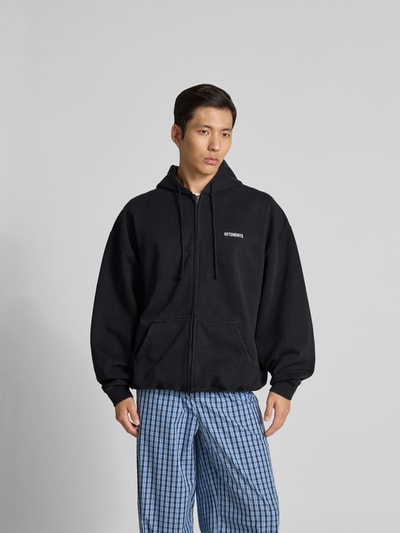 Männermodel trägt Oversized Sweatjacke aus Baumwolle von VETEMENTS in Schwarz mit Zweiwege-Reißverschluss, Känguru-Tasche, elastische Abschlüsse, Ganzkörperansicht von vorne. Rückenlänge 75 cm, Schulterbreite 50 cm, Ärmellänge 78 cm.