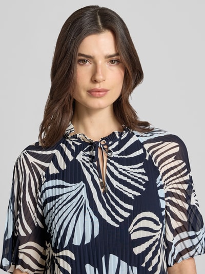 Modelka nosi Regular Fit Blusenshirt in Plissee-Optik od s.Oliver BLACK LABEL, efekt: wzór na całej powierzchni, krój regular fit, kształt kołnierza: kołnierz z falbanami, detale: plisy, dekolt: okrągły dekolt, wycięcie w kształcie łezki, zapięcie: wiązanie, rękaw: krótki rękaw, widok: z przodu, zbliżenie na górną część ciała.