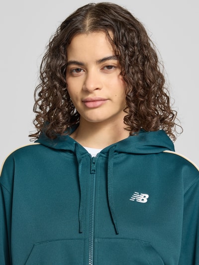 Frauenmodel trägt Cropped Sweatjacke mit Label-Stitching von New Balance in Dunkelgrün, Kapuze mit Tunnelzug, Reißverschluss, Känguru-Tasche, Nahaufnahme Oberkörper.