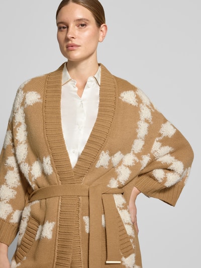 Frauenmodel trägt Cardigan mit Bindegürtel und 3/4-Arm von Marc Cain, beiger Allover-Look mit weißem Muster, Material-Mix mit Alpaka-Anteil, Nahaufnahme Oberkörper.