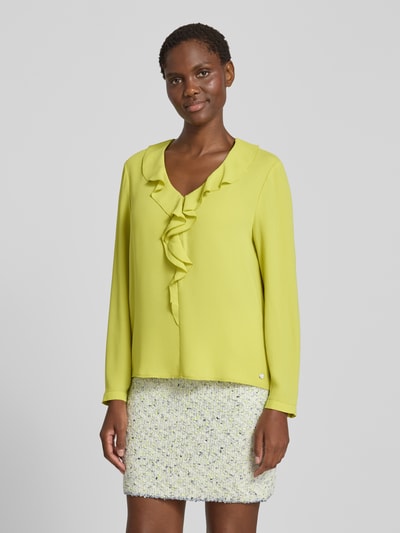 Kobiece model prezentuje Relaxed Fit Bluse mit Rüschenbesatz od Marc Cain, bluzka z obszyciem falbanami, dekoltem w serek, z długim rękawem, długość tyłu w rozmiarze 36: 58 cm, długość rękawów w rozmiarze 36: 61 cm, jasny żółty kolor, gładka tekstura, widok całej sylwetki z przodu.