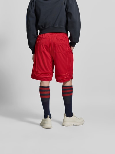 Männermodel trägt Baggy Fit Sweatshorts mit Tunnelzug von RHUDE in Rot mit Mesh, Label-Stitching, Eingrifftaschen, elastischer Bund, Ganzkörperansicht Rückansicht.