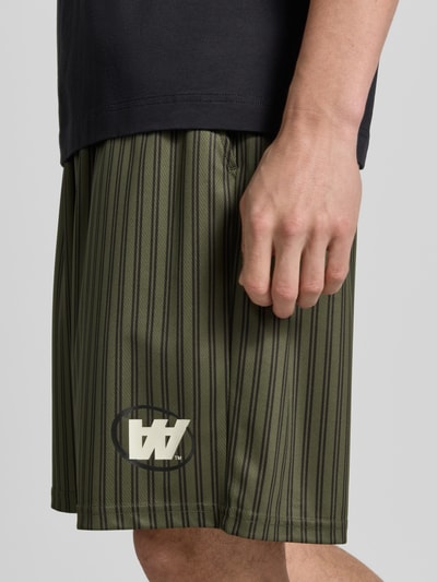 Männermodel trägt Regular Fit Shorts mit Label-Print Modell 'Lama' von Wood Wood, grün mit schwarzen Streifen, Nahaufnahme Oberschenkel, elastischer Bund, Kontext: Herren Shorts aus Regular Fit, elastischer Bund, Label-Print, Bundweite bei Größe M: 40 cm, Innenbeinlänge bei Größe M: 19 cm.