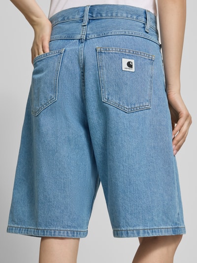 Frauenmodel trägt Relaxed Fit Jeans Shorts aus reiner Baumwolle Modell 'Brandon' von Carhartt Work In Progress, hellblauer Denim, 5-Pocket-Design, Label-Patch, Knopf-Reißverschluss, Ganzkörperansicht Rückansicht.
