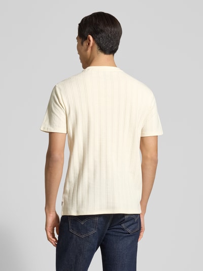 Męski model nosi Regular Fit T-Shirt aus Baumwolle od Guess, kremowy kolor, faktura prążków, detal z logo, długość tyłu 69 cm, krój regular fit, okrągły dekolt, widok całej sylwetki od tyłu.