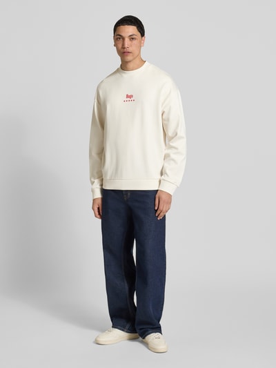Männermodel trägt Regular Fit Sweatshirt mit Happy Hugo Print von Hugo Blue in Creme mit rotem Print, reine Baumwolle, Vorderansicht Ganzkörperansicht.