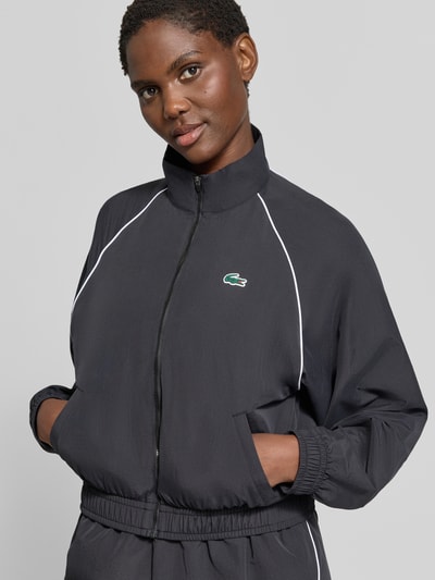 Frauenmodel trägt Relaxed Fit Trainingsjacke mit Logo-Patch von Lacoste in Schwarz mit weißem Piping, glatte Struktur, Nahaufnahme Oberkörper, Damen Trainingsjacke von Lacoste Sport.