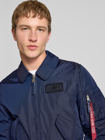 Männermodel trägt Regular Fit Bomberjacke mit Logo-Patch von Alpha Industries in Dunkelblau, Umlegekragen, Nahaufnahme Oberkörper. Rückenlänge 66 cm, Schulterbreite 44 cm, Brustweite 56 cm, Ärmellänge 66 cm, mit Reißverschluss, AufgesetzteTaschen, elastische Abschlüsse.