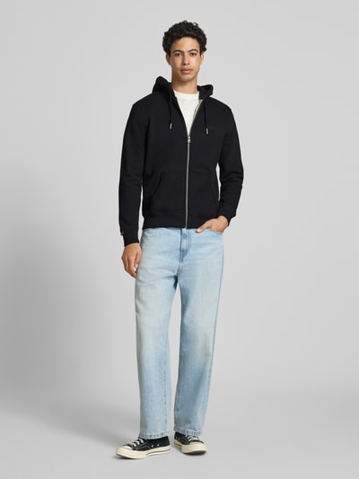 Männermodel trägt Sweatjacke mit Kapuze und Logo-Stitching von Superdry, Baumwoll-Mix, Regular-Fit, Schwarz, Ganzkörperansicht von vorne. Rückenlänge bei Größe M: 66 cm, Ärmellänge bei Größe M: 62 cm, Kapuze mit Kordelzug, Reißverschluss, Seitliche Eingrifftaschen, Gerippte Abschlüsse, Label-Detail