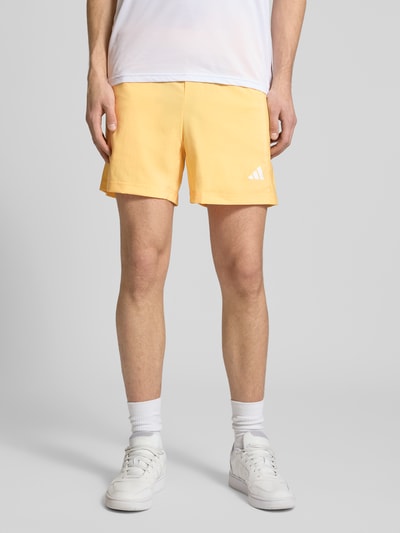 Męski model nosi Regular Fit Sport-Shorts mit Label Print von adidas Training, efekt: jednolity kolor, krój regular fit, nadruk z logo, elastyczny ściągacz z tunelem, kieszenie zapinane na zamek błyskawiczny, Ganzkörperansicht von vorne.