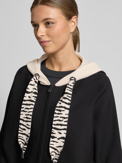 Vrouwenmodel draagt Strickjacke mit Reißverschluss van Marc Cain in zwart met beige capuchon, viscose-mix, losse pasvorm, 3/4-mouw, detailopname bovenlichaam.
