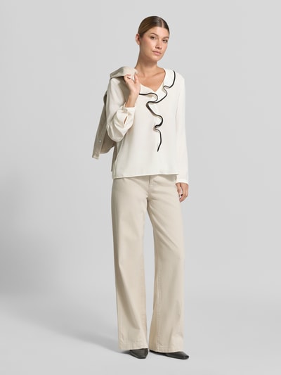 Frauenmodel trägt Regular Fit Bluse mit Volants von Marc Cain in Creme mit schwarzem Rand, V-Ausschnitt, Label-Detail, fließende Textur, Ganzkörperansicht von vorne.