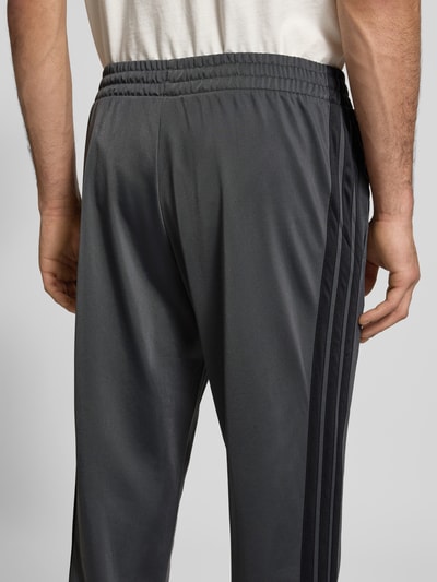 Männermodel trägt Slim Fit Trainingsanzug mit Logo-Stitching von adidas Sportswear in Dunkelgrau mit schwarzen Seitenstreifen, Detailaufnahme Rückenansicht. Bundweite, Rückenlänge, Schulterbreite, Ärmellänge und Innenbeinlänge laut Kontext.