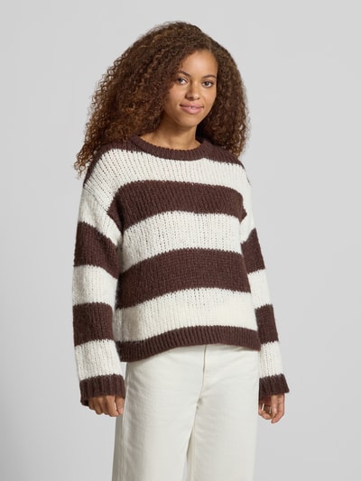 Modelka kobieca nosi sweter Relaxed Fit Pullover im Chunky Knit Look model 'DORTHEA' od PIECES, szerokie brązowo-białe paski, dzianina o wyraźnej fakturze, dziewczęcy styl, Ganzkörperansicht von vorne.