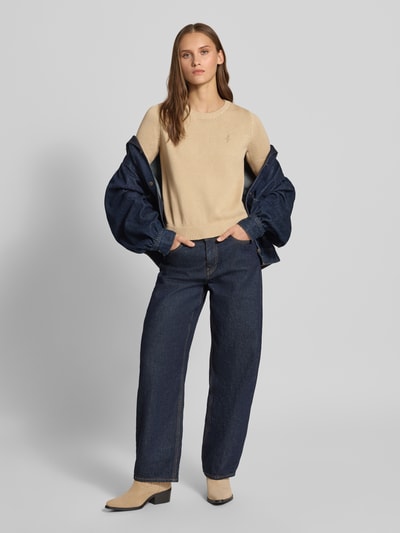 Frauenmodel trägt Slim Fit Pullover aus reiner Baumwolle von REVIEW in Strick-Optik, beige, kombiniert mit dunkler Jeans und beigen Schuhen, Ganzkörperansicht von vorne. Rückenlänge 54 cm, Ärmellänge 72 cm, Logo-Stitching.