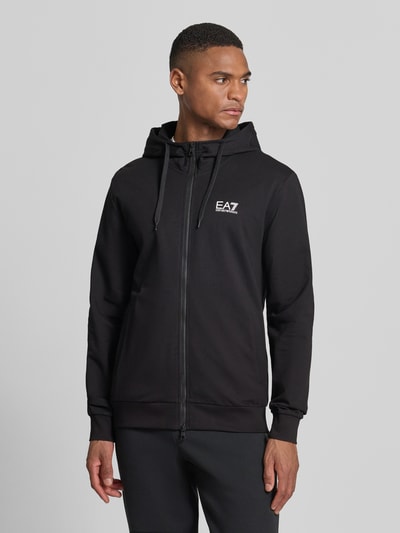 Męski model nosi Slim Fit Sweatjacke mit Kapuze od EA7 Emporio Armani, czarny kolor, mieszanka bawełny, nadruk z logo, wpuszczane kieszenie, elastische wykończenia, zamek błyskawiczny dwustronny, kaptur z tunel, stójka, długość tyłu 69 cm, długość rękawów 69 cm, krój slim fit, Ganzkörperansicht z przodu.