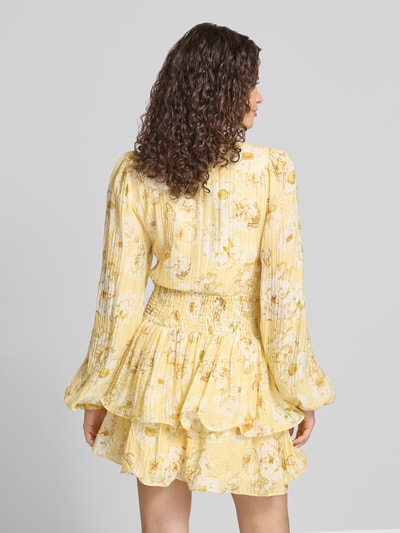 Frauenmodel trägt Minikleid aus Viskose-Mix mit Rüschenbesatz von Gina Tricot in Gelb mit floralen Mustern, taillierter Passform und langen Ärmeln, Rückenlänge 80 cm, Brustweite 41 cm, Ärmellänge 69 cm, Ganzkörperansicht Rückansicht.