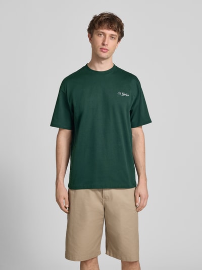 Männermodel trägt Loose Fit T-Shirt aus reiner Baumwolle Modell 'LES CLASSIQUES' von Only & Sons, Optik: Unifarben, Passform: Loose-Fit, Applikation: Schriftzug, Ausschnitt: Rundhals, Ärmelform: Kurzarm, Materialverarbeitung: Jersey, dunkles Grün, glatte Textur, Ganzkörperansicht von vorne.
