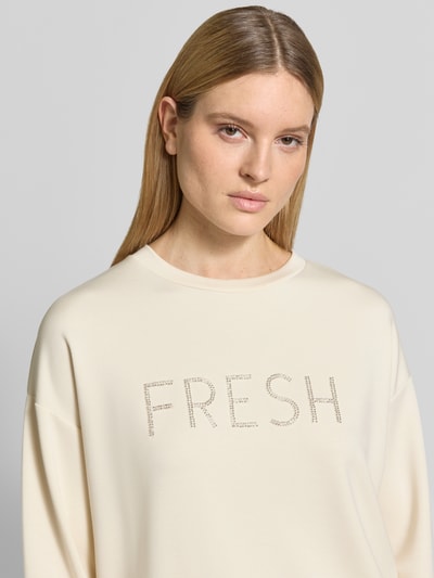 Modelka kobieca ma na sobie Regular Fit Sweatshirt mit Ziersteinbesatz Modell 'Banu' od Soyaconcept, kremowy kolor, gładka faktura, okrągły dekolt, elastyczne wykończenia, detal z cyrkoniami na przodzie, mieszanka materiałów z dodatkiem modalu, krój regular fit, zbliżenie na górną część ciała.