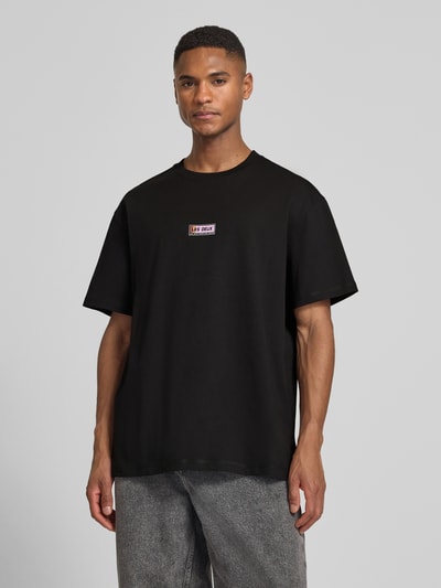 Męski model nosi T-Shirt mit Logo-Print Modell 'Chase' von Les Deux z czystej bawełny, prosty krój, okrągły dekolt, widok z przodu, ujęcie całej sylwetki.