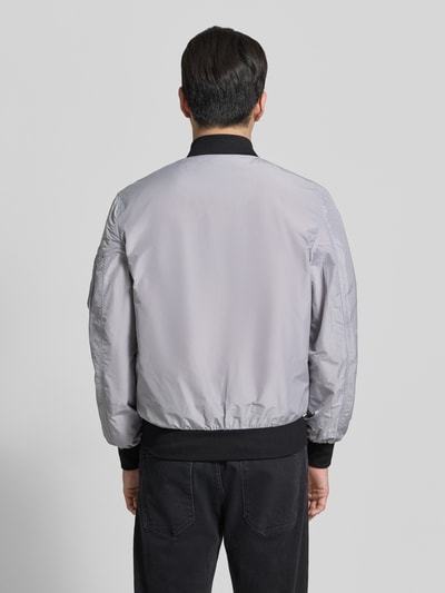 Männermodel trägt Bomberjacke mit Label-Patch und Eingrifftaschen von Original Bombers, grauer Farbton, Gerade geschnitten, Rückenansicht, Kontext: Herren Bomberjacke, Reißverschluss, Reißverschlusstasche am Ärmel, Druckknopfverschluss, Gerippte-Abschlüsse, Rückenlänge Größe M 67 cm, Schulterbreite 43 cm, Brustweite 53 cm, Ärmellänge 68 cm.