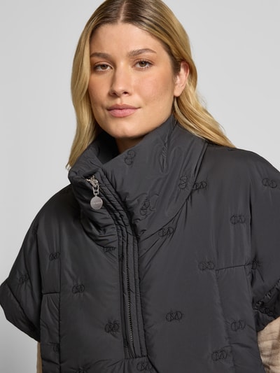 Vrouwenmodel draagt Cape met Reißverschluss und Eingrifftaschen Modell 'NETTI' von Cph Muse in zwart met lichte structuur; regular fit, opstaande kraag, detailopname bovenlichaam, ruglengte bij maat S/M 61 cm, borstwijdte bij maat S/M 64 cm.