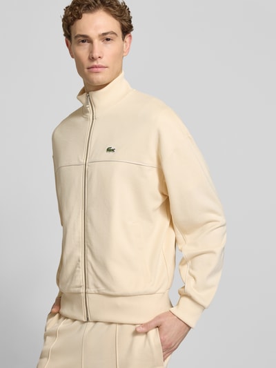 Męski model nosi Regular Fit Sweatjacke mit Baumwoll-Anteil od Lacoste, efekt: jednolity kolor, krój regular fit, zamek błyskawiczny, detal z logo, paski w kontrastowym kolorze, szew z wypustkami, stójka, długi rękaw, prążkowane zakończenia, wpuszczane kieszenie, dzianina dresowa, mieszanka materiałów z bawełną, Ganzkörperansicht von vorne.