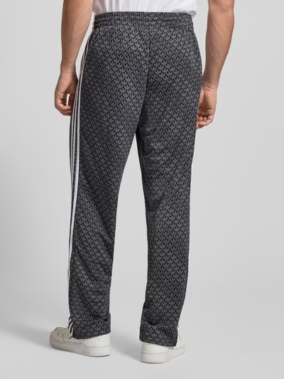 Männermodel trägt Trainingshose mit Logo-Muster von adidas Originals, Relaxed-Fit, Label-Stitching, elastischer Bund, Ganzkörperansicht Rückansicht.