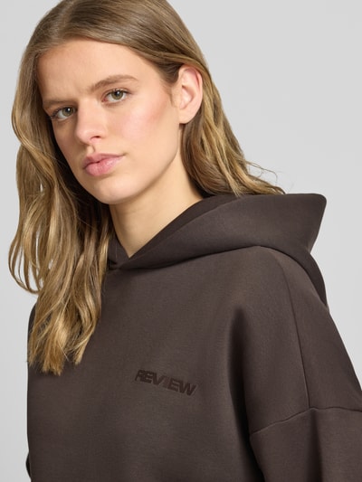 Modelka nosi Hoodie mit Logo und Känguru-Tasche von Review aus mieszanka bawełny w kroju relaxed fit, kolor brązowy, logo, kieszeń kangurka. Zbliżenie na górną część ciała.