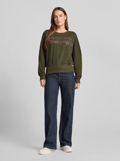 Kobiece modelka nosi Oversized Sweatshirt aus reiner Baumwolle von miss goodlife, krój oversized, bawełna, okrągły dekolt, nadruk z napisem, wykończenia: elastyczne, kolor oliwkowy, gładka faktura, pełny przód, jeansy, białe buty, całkowity widok z przodu.