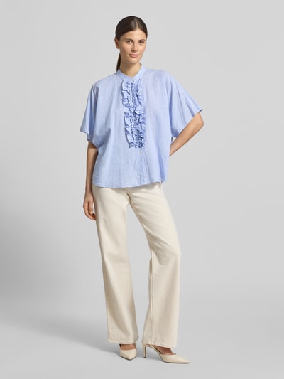 Modelka kobieca nosi Relaxed Fit Bluse mit Rüschen Modell 'DAISY' od 0039 Italy, len-bawełna, jasnoniebieska, prosty fason, stójka, kremowe spodnie, Widok całej sylwetki z przodu.