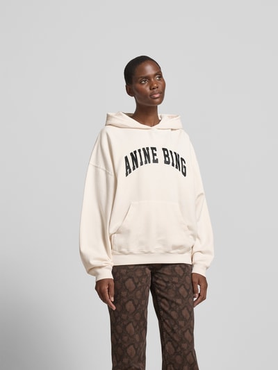 Frauenmodel trägt Oversized Hoodie mit Logo-Print von Anine Bing aus Reiner Baumwolle, Oversized-Fit, Logo-Print, Känguru-Tasche, elastische Abschlüsse, Rückenlänge bei Größe S 68 cm, helle Farbe, lockere Form, weiche Textur. Ganzkörperansicht von vorne.