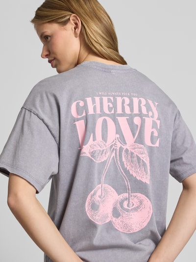 Frauenmodel trägt Oversized T-Shirt aus Bio-Baumwolle Modell 'KINNA LIFE' von Only, z efektem melanżu, krój oversized, aplikacja z napisem, okrągły dekolt, krótki rękaw, zrolowany brzeg, materiał: czysta bawełna ekologiczna, kolor szary z różowym nadrukiem, Ganzkörperansicht Rückansicht.
