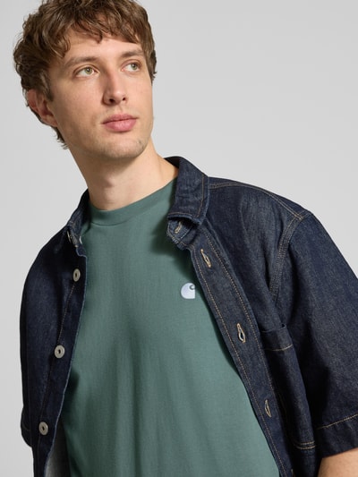 Męski model nosi T-Shirt mit Label-Stitching Modell 'Madison' od Carhartt Work In Progress, czysta bawełna, krój regular fit, zielony kolor, okrągły dekolt, detal haftowanego logo, długość tyłu w rozmiarze M: 75 cm, zbliżenie na górną część ciała.