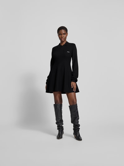 Frauenmodel trägt Strickkleid mit Label-Detail von ROTATE aus Modal-Woll-Mix, taillierter Schnitt, Hemdblusenkragen, schwarze Farbe, Ganzkörperansicht von vorne.
