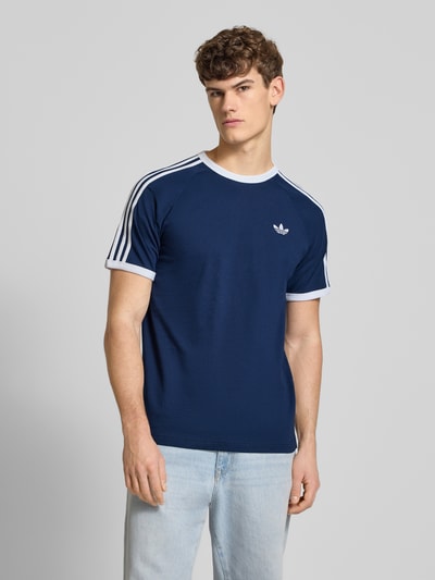 Männermodel trägt T-Shirt mit Label-Stitching von adidas Originals in Dunkelblau mit weißen gerippten Abschlüssen und Rundhalsausschnitt aus reiner Baumwolle, Regular-Fit, Rückenlänge bei Größe M 73 cm, Ganzkörperansicht von vorne.