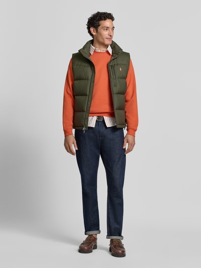 Männermodel trägt Regular Fit Weste mit Logo-Stitching von Polo Ralph Lauren in Oliv, kombiniert mit orangenem Pullover, Ganzkörperansicht von vorne.