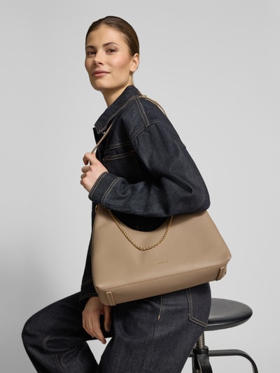Modelka nosi Handtasche mit Label-Detail od CK Calvin Klein w beżowym kolorze, skórzana tekstura, łańcuszkowy pasek na ramię, Szerokość w rozmiarze 1: 24 cm, długość 27 cm, głębokość 11 cm, długość paska 68 cm, zapięcie na zamek, kieszeń wewnętrzna na zamek, widok całej sylwetki z przodu.