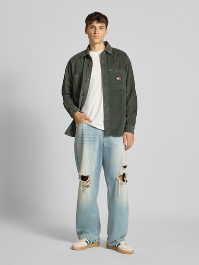 Männermodel trägt Relaxed Fit Cordhemd aus reiner Baumwolle von Tommy Jeans in Dunkelgrün mit Kentkragen, aufgesetzter Brusttasche und Logo, Ganzkörperansicht von vorne