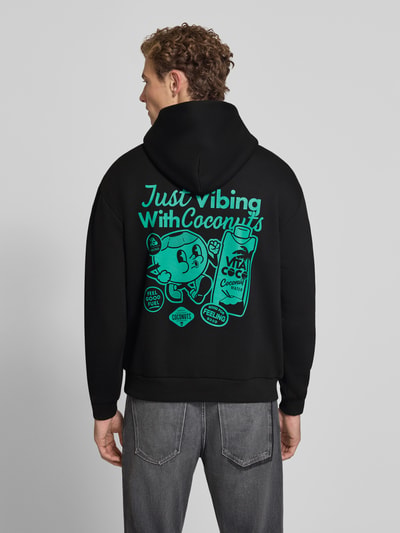 Männermodel trägt Relaxed Fit Hoodie mit Motiv-Print von REVIEW x ANSON´S, Baumwoll-Mix, schwarze Farbe mit türkisem Print, Ganzkörperansicht Rückansicht.