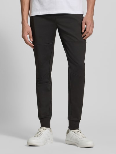 Męskimodel nosi spodnie dresowe z elastycznej mieszanki materiałów z mieszanką bawełny model 'GORDON' od Jack & Jones, krój slim fit, elastyczny pas z tunelem, elastyczne zakończenia nogawek, boczne kieszenie zapinane na zamek błyskawiczny, długość wewnętrzna nogawki w rozmiarze M: 73 cm, kolor czarny, gładka tekstura, przód, ujęcie całej sylwetki