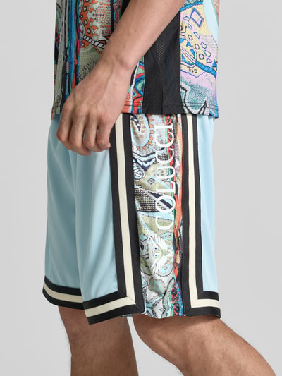 Mężczyzna nosi Shorts aus Mesh mit Kontraststreifen od COLUCCI w jasnoniebieskim kolorze, z elastycznym paskiem i kontrastowymi pasami, szerokość w rozmiarze M: 37 cm, długość nogawki od wewnątrz: 24 cm, ujęcie zbliżeniowe dolnej części ciała.