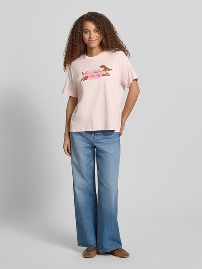 Frauenmodel trägt Regular Fit T-Shirt aus reiner Baumwolle Modell 'VIVVI' von Only, Optik: Unifarben, Motiv-Print, Rundhals, Kurzarm, Materialverarbeitung: Jersey, Material: Baumwolle-Mix, sanftes Rosa, lockere Form, glatte Textur, Ganzkörperansicht von vorne.