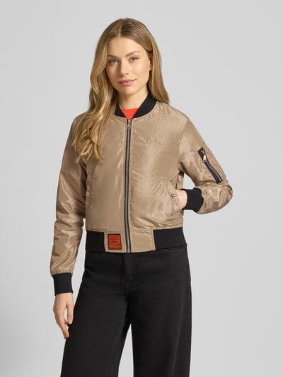 Frauenmodel trägt Bomberjacke mit Label-Patch und Reißverschlusstasche am Ärmel von Original Bombers in Beige mit schwarzen elastischen Abschlüssen, Logo-Stitching, Stehkragen, seitliche Eingrifftaschen mit Druckknopfverschluss; Ganzkörperansicht von vorne. Rückenlänge 53 cm, Schulterbreite 37 cm, Brustweite 48 cm, Ärmellänge 63 cm.