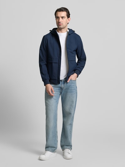 Männermodel trägt Regular Fit Funktionsjacke mit Kapuze von Tom Tailor Denim in Dunkelblau, unifarben, elastische Abschlüsse, Ganzkörperansicht von vorne. Die Jacke ist wasserdicht, atmungsaktiv und wärmeisolierend.