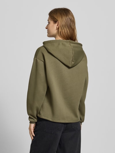 Vrouwenmodel draagt Regular Fit Hoodie uit katoenmix model 'CHILLI' van Pieces in effen olijfgroen sweatmateriaal, merklabel detail aan de achterkant, geribde manchetten, lange mouwen, rugaanzicht.