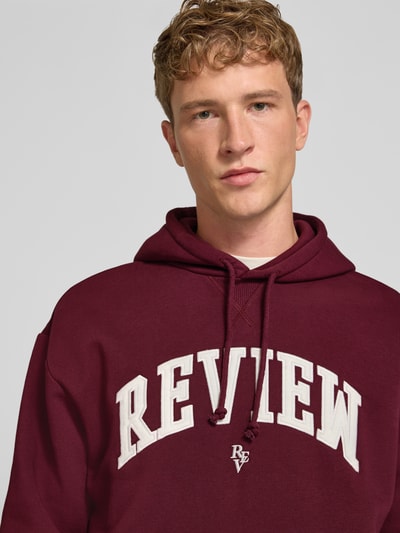 Męskimodel nosi Relaxed Fit Hoodie aus Baumwoll-Mix mit Logo-Stickerei od REVIEW, mieszanka bawełny, krój relaxed fit, wyhaftowane logo, kaptur z tunelem, krój z długim rękawem, prążkowane wykończenia, kieszeń kangurka, długość tyłu w rozmiarze M: 67 cm, długość rękawów w rozmiarze M: 65 cm, kolor bordowy, zbliżenie na górną część ciała.