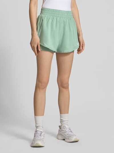 Frauenmodel trägt Shorts im 2-in-1-Look mit Logo von NIKE TRAINING in Mintgrün, elastischer Bund, Regular-Fit, Ganzkörperansicht von vorne.