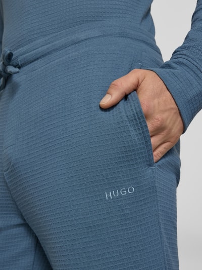 Mężczyzna nosi Relaxed Fit Pyjama-Hose aus Baumwoll-Mix Modell 'AUSTIN PANTS' von HUGO w kolorze niebieskim, faktura waflowa, nadruk z logo, widok z bliska na kieszeń.